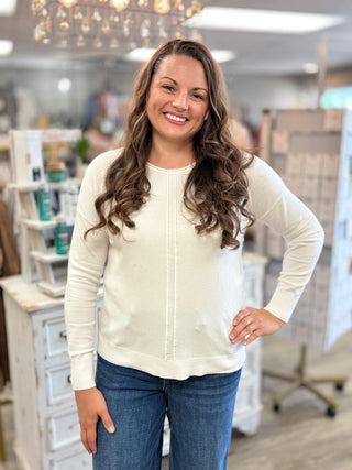 A smiling woman with long wavy brown hair stands in a retail store wearing a cream-colored long-sleeve knit sweater and blue jeans.