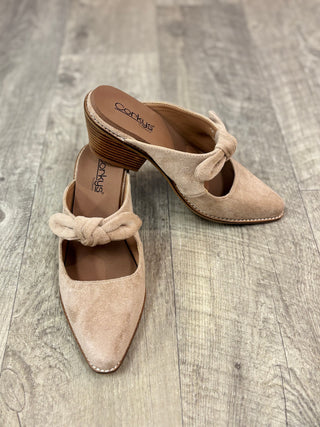 A pair of beige suede mule heels with pointed toes and knotted bow details on the instep, displayed on a gray wood-plank floor. 