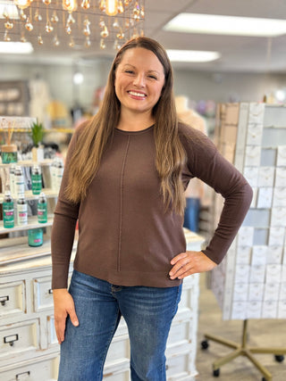 A woman with long light brown hair stands in a retail store wearing a brown long-sleeve knit top with a vertical seam detail down the center and blue jeans.