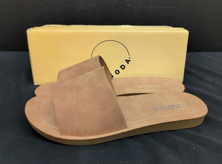 Brown sandal with 'Soda’ branding on a black surface, next to a yellow box with the brand logo.