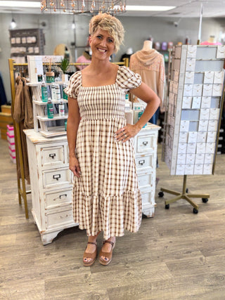 A woman modeling a beige and brown gingham midi dress with puff sleeves and a smocked bodice, standing in a boutique setting.