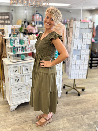 A woman modeling an olive green tiered maxi dress in a retail store setting, standing next to white display cabinets.