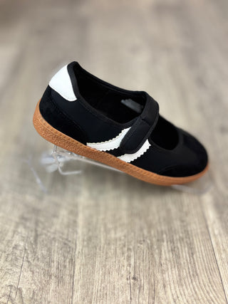 A single black Mary Jane shoe with a white side stripe and a brown gum sole, displayed on a clear plastic stand over a light wood floor.