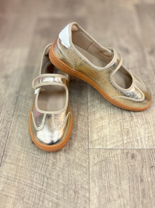 A pair of metallic gold Mary Jane shoes with tan soles, displayed on a light wood floor.