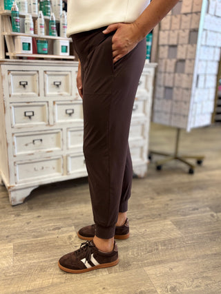 A person wearing dark brown jogger pants and matching brown sneakers stands in a retail store with a white cabinet and wood floor in the background.