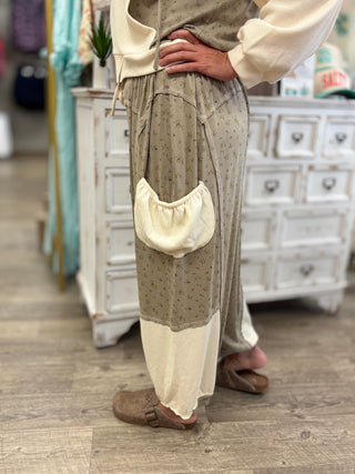 A person wearing a bohemian-style two-piece outfit with a beige and cream color-blocked skirt featuring a small floral print and a large cream pocket, paired with a cream long-sleeve top and brown clogs, in a rustic shop setting.