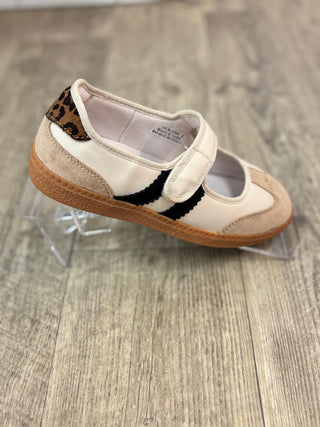 A Mary Jane shoe in off-white leather with a black scalloped side stripe, a leopard-print heel tab, and a brown gum sole, displayed on clear acrylic stands over a gray wood-look floor.