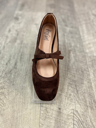 A dark brown suede Mary Jane flat shoe with a bow detail across the instep, displayed on a clear acrylic stand over a gray wood-look floor.