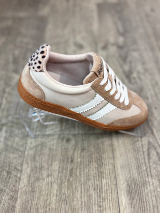 A low-top sneaker with beige suede panels, white leather overlays, white laces, a brown rubber sole, and a black-and-white spotted pattern on the heel tab, displayed on a clear stand over a gray wood floor.