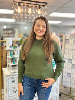A woman with long brown hair wearing an olive green knit sweater and blue jeans, standing in a retail store setting with a white cabinet and hanging lights in the background.