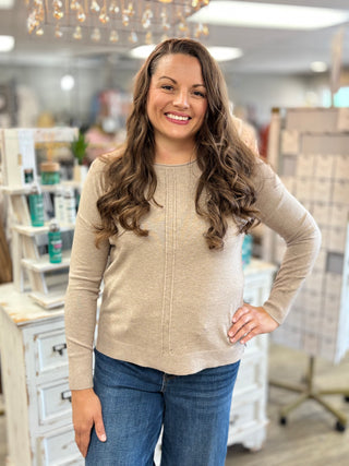 A smiling woman with long wavy brown hair stands in a boutique wearing a beige knit long-sleeve top and blue jeans.