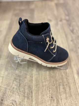 A single navy blue suede ankle boot with beige laces and a cream sole, displayed on a clear plastic stand over a light wood floor.