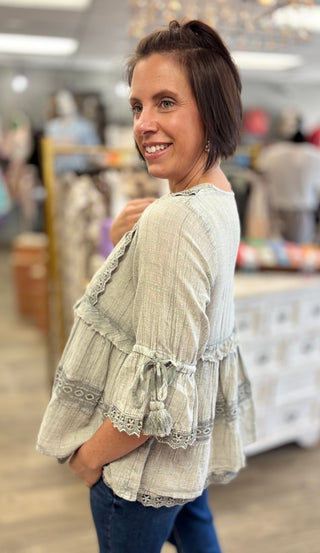 A woman modeling a light sage green gauze blouse with ruffled sleeves and lace trim, standing in a clothing store.