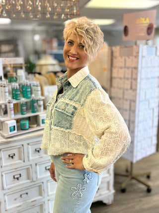 A woman with short blonde hair stands in a boutique shop wearing a light blue denim jacket with a cream lace overlay and light blue jeans.