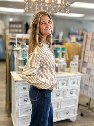 A woman with blonde hair smiling in a boutique, wearing a beige and white striped long-sleeve knit top and dark blue jeans.
