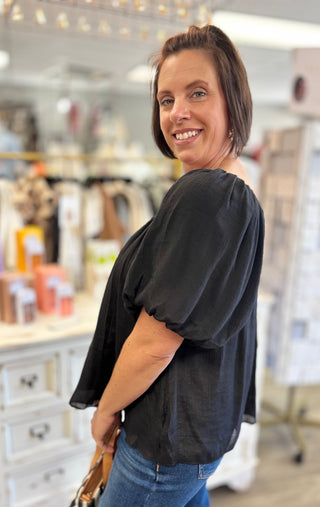 A woman with short brown hair smiling and looking back over her shoulder while wearing a black puff-sleeve blouse and blue jeans inside a retail store.