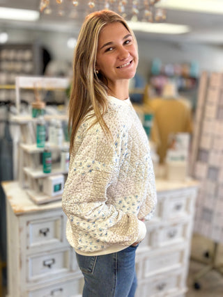 A smiling young woman with long blonde hair stands in a boutique shop, wearing a cream-colored textured knit sweater with a small blue floral pattern and blue jeans.