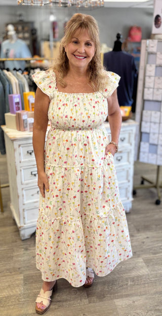 Model wearing long cream dress with floral print, flutter sleeves, smocked bodice, and pockets