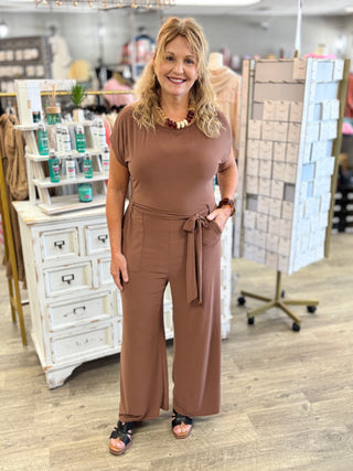 A woman modeling a brown short-sleeve jumpsuit with wide legs and a tie waist, standing in a retail store setting.