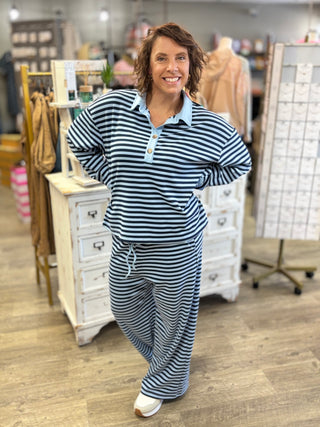 A woman modeling a matching two-piece loungewear set with navy and light blue horizontal stripes, featuring a long-sleeve polo shirt with a light blue collar and matching drawstring pants, set in a retail store.