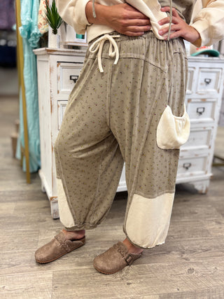 A person wearing loose, draped beige pants with a small floral pattern, cream-colored contrast panels at the ankles, a drawstring waist, and a side pouch pocket. The person is standing in a rustic shop setting with white wooden furniture and a wood floor.