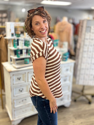A woman wearing a brown and cream striped short-sleeve knit top and blue jeans, standing in a retail store.