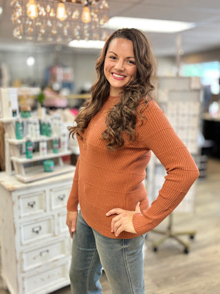 A smiling woman with long wavy brown hair stands in a retail store wearing a ribbed knit long-sleeve sweater in a warm rust orange color, paired with light blue jeans.