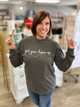 A woman smiling in a shop setting, wearing a dark gray long-sleeve t-shirt with the phrase 'get your hopes up.' printed in white script across the chest, paired with blue jeans.