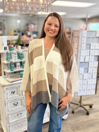 A smiling woman stands in a retail store wearing a cream, beige, and taupe open-front cardigan over a beige top and blue jeans.
