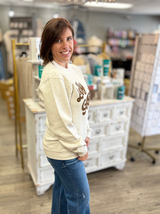 A woman with short brown hair smiling and posing in a boutique, wearing a cream-colored crewneck sweatshirt with a brown graphic print and blue jeans.