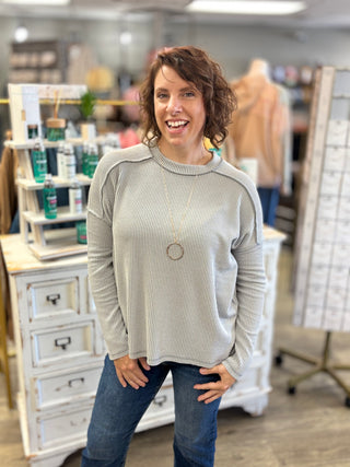 A woman with curly brown hair smiling in a retail store, wearing a light gray ribbed long-sleeve sweater and blue jeans.