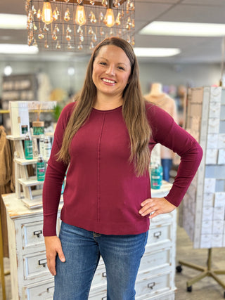 A woman with long brown hair stands in a retail store wearing a long-sleeve crewneck top in deep burgundy and blue jeans.