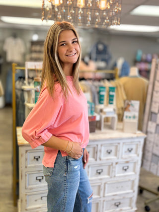 A young woman smiling in a boutique, wearing a coral pink long-sleeve top and light wash blue jeans, standing in front of a white wooden dresser.