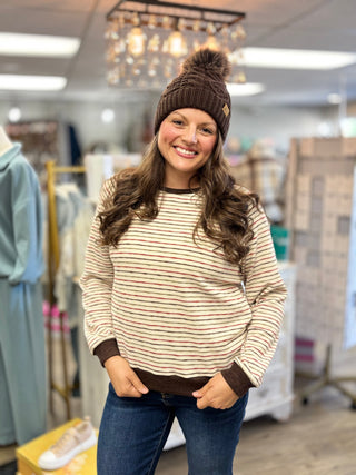 Front view of a dark brown cable knit pom hat with a folded ribbed cuff, paired with a cream striped top.