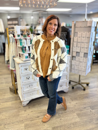 Model wearing a camel colored suede top with snap and ruffle detail, a cream argyle printed cardigan, jeans, and loafers.