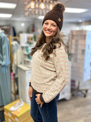 Angled view of a dark brown cable knit hat showing its fluffy pom and cuff patch, worn with a striped top.