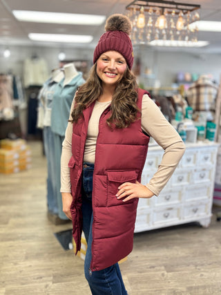 Burgundy cable knit pom hat styled with a matching long puffer vest, beige long-sleeve top and blue jeans.