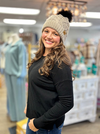 Angled view of a light taupe cable knit hat with a dark pom, ribbed cuff and small patch, worn with a black sweater.