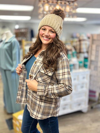 Angled view of a tan cable knit pom hat with a ribbed cuff and small patch, paired with a plaid shirt jacket.